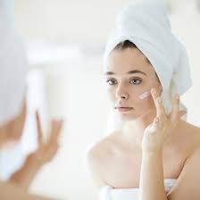 A close-up of a young person applying for navigating acne treatment cream to their cheek in front of a bathroom mirror.